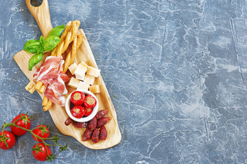 Italian appetizers (antipasti) on concrete background with copy space. Prosciuto, grissini, pickled peppers and salami on wooden board, fresh basil and tomato. 