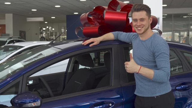 Attractive male customer showing his thumb up near the car at the showroom. Handsome caucasian guy approving car dealership. Young man leaning his hand on purple hatchback with gift bow on it's roof
