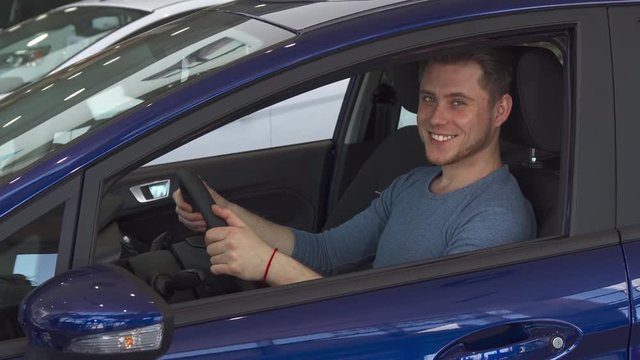 Handsome male customer smiling for the camera from inside the car. Attractive caucasian sitting inside the purple hatchback. Young man touching the steering wheel of the vehicle at the dealership