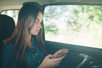 Beautiful woman sitting on the back passenger seats in the car. Girl is using a smartphone