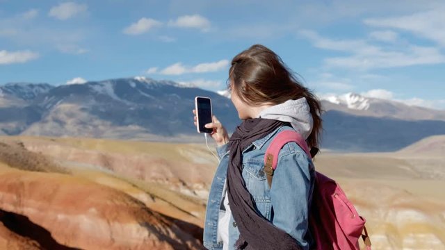 Attractive sporty 20s woman in a denim jacket and scarf takes pictures of mountains on the smartphone 20s 4k.