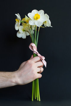 Lovely Bouquet Of White And Yellow Narcissus With A Bow Made Of  Pink Ribbon In The Hand Of A Married Man On A Black Background. Nice For Postcard