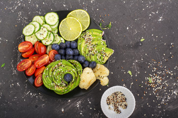 A healthy snack from tomatoes, cucumbers, cauliflower, avocado and blueberries with slices of lime and sesame seeds in a black plate on a dark background. Top View