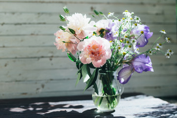 Bouquet of white peonies, chamomiles and iris flowers in glass vase. Summer background. Tinted photo