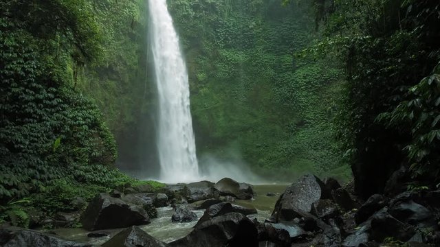 Amazing Nungnung waterfall, slow motioned falling water hitting water surface, some huge rocks seeable in front of frame. Lush green leafes is moving from the wind, Bali, Indonesia