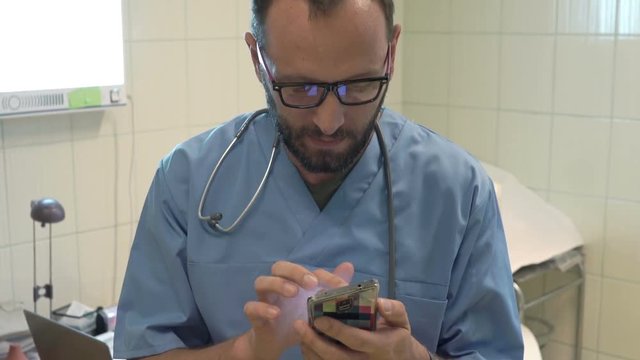 Young, male doctor browsing Internet on smartphone sitting by table in office

