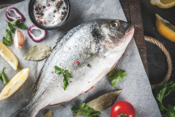 Fresh raw organic fish Dorado, sea bream with ingredients for cooking lemon, tomatoes, onion, salt, pepper, bay leaf, greens, oil. On old wooden cutting board, on black stone table Top view copy space