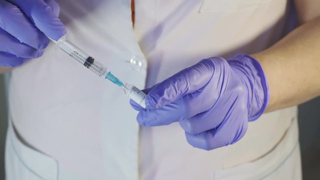 Young nurse fills disposable syringe with medicine indoors. She lowers long metal needle into glass ampoule and slowly pulls plastic piston of injector to fill him with liquid preparation to mark that