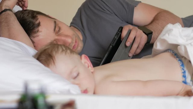 Father reads the news on the tablet near the sleeping baby. A boy resting in the foreground of a house on a bed