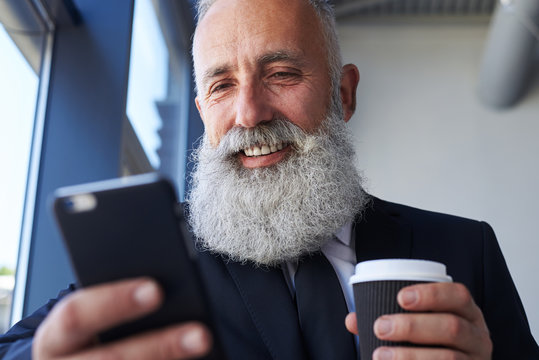 Pleased Gentleman Age Of 50-60 Holding Cup Of Coffee And Surfing In Phone