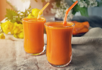 Carrot juice into two glass cups on a background of carrots and flowers in the early morning. The horizontal frame.