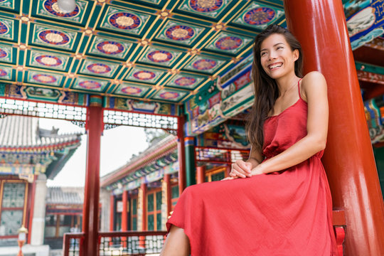 Beautiful asian young woman relaxing on summer holiday by ancient traditional wooden red door in Chinese temple on china travel destination. Girl tourist in elegant silk sun dress.