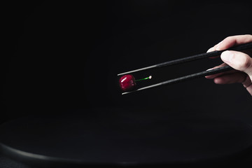 Hand holding black chopsticks with a red cherry on a black background.