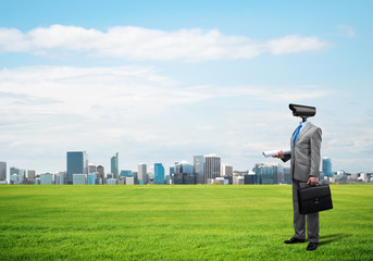 Camera headed man standing on green grass against modern cityscape