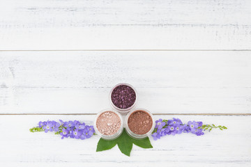 Glitter powder make up with purple flowers and green leaves on white wood background with copy space
