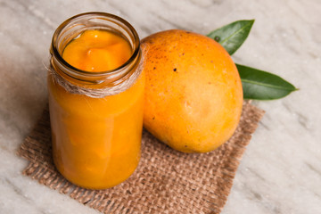 Homemade Mango Jam with fresh fruits and leaves of mango on a white marble.