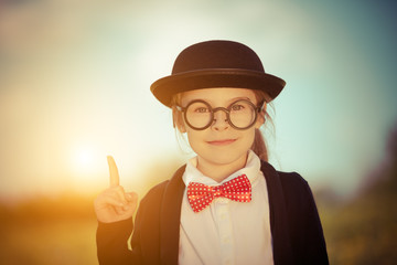 Funny little girl in glasses, bow tie and bowler hat pointing finger up. Retro stile.