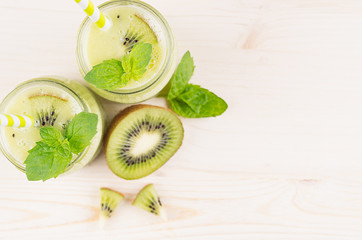 Fototapeta na wymiar Green kiwi fruit smoothie in glass jars with straw, mint leaf, cute ripe berry, top view. White wooden board background, decorative border, copy space.
