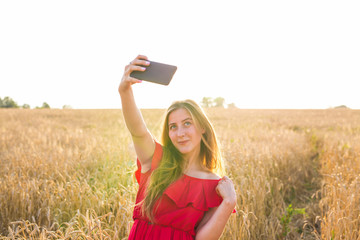 technology, summer holidays, vacation and people concept - smiling young woman in red dress taking selfie by smartphone on cereal field