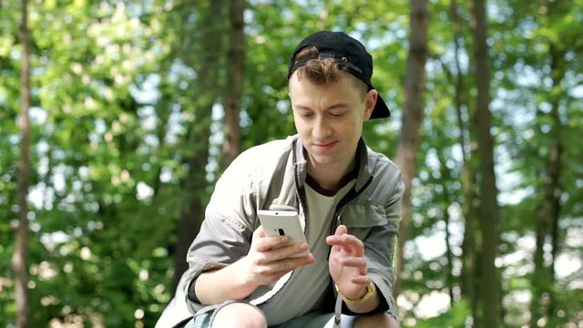 Stylish boy sitting in the park and doing selfies on smartphone, steadycam shot
