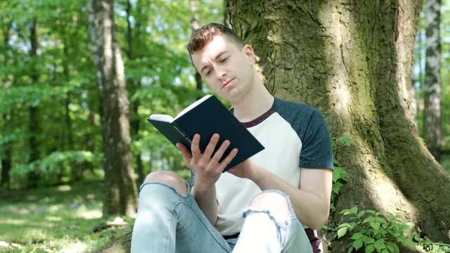 Young man reading book in the park and smiling to the camera, steadycam shot
