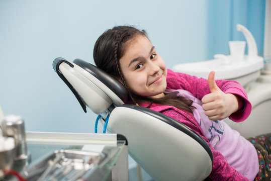 Happy patient girl showing thumbs up at dental office. Medicine, stomatology and health care concept. Dental equipment
