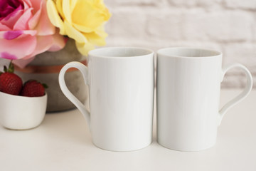 Fototapeta na wymiar Two Mugs. White Mugs Mockup. Blank White Coffee Mug Mock Up. Styled Photography. Coffee Cup Product Display. Two Coffee Mugs On White Desk. Vase With Pink Roses
