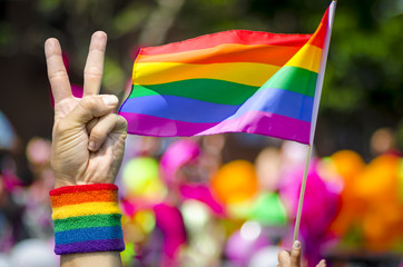 Hand wearing gay pride rainbow flag wristband holding up a victory/peace sign gesture in front of...