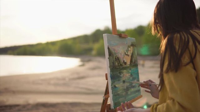Mysterious girl finishes painting still life with oil paints, the artist went to the sea plein air, she draws the sea or river and mountain with the help of mixing flowers