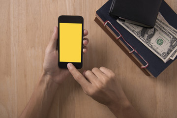Top view of hand holding smartphone with yellow blank screen on wood desktop.