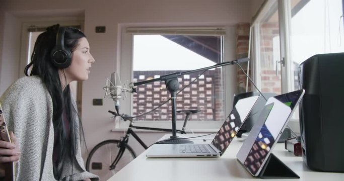 woman creative musician at home studio works by playing,singing and recording guitar with notebook tablet and microphone indoor in modern industrial house. caucasian brunette. 4k handheld side video