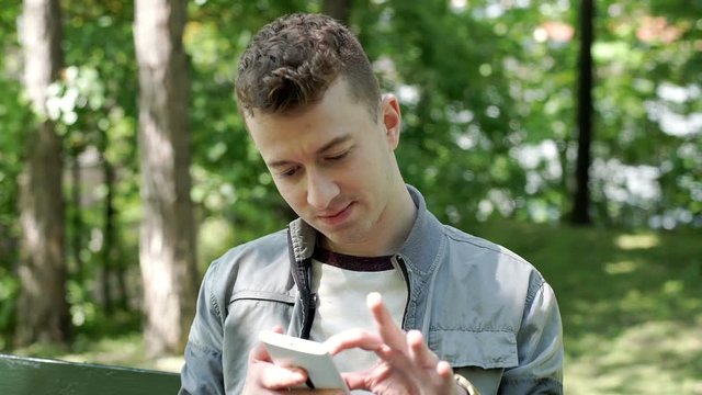 Young man sitting in the park and browsing internet on smartphone, steadycam shot
