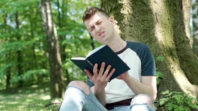 Young man reading book in the park and having painful headache, steadycam shot
