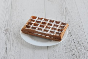 Chocolate waffle with powdered sugar on a white plate on a wooden table