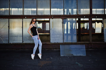 Girl at sunglasses and jeans posed at the roof of abadoned industrial place with windows.