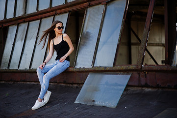 Girl at sunglasses and jeans posed at the roof of abadoned industrial place with windows.