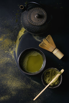 Green tea matcha powder and hot drink in black bowls standing with iron teapot, bamboo traditional tools spoon and whisk over dark metal background. Top view with space
