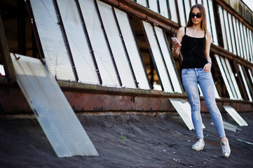 Girl at sunglasses and jeans, with phone and headphones posed at the roof of abadoned industrial place with windows.