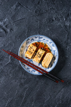 Japanese Rolled Omelette Tamagoyaki sliced with black sesame seeds and soy sauce, served in blue white ornate ceramic plate with chopsticks over black stone texture background. Top view with space
