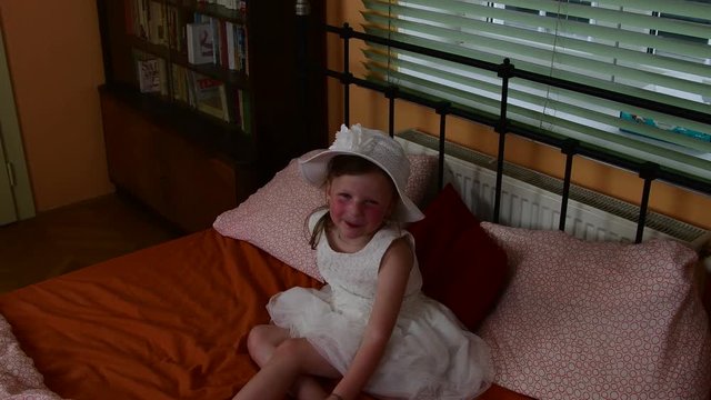 A cute small girl is sitting and smiling on the bed. Parents bedroom and  her daughter is on the bed.  A small girl is wearing white dress.