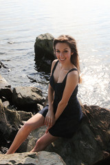 A sexy young model smiles, sits on rocks by ocean.