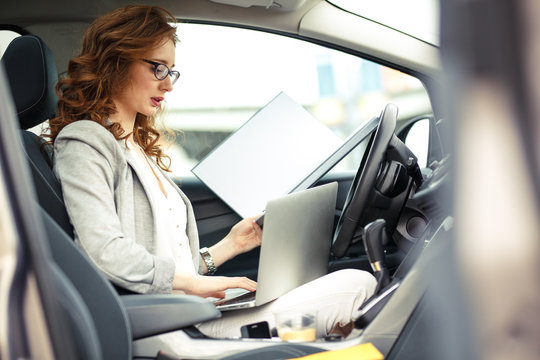 Attractive female sales manager using laptop and phone in the car.Preparing and looking into contract papers.