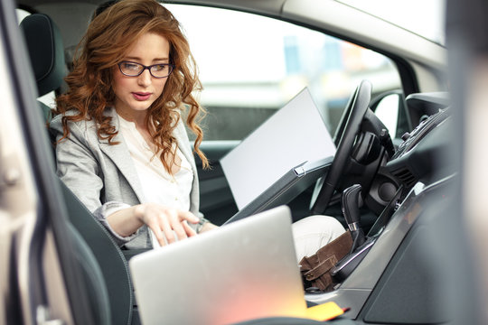 Attractive female sales manager using laptop and phone in the car.Preparing and looking into contract papers.