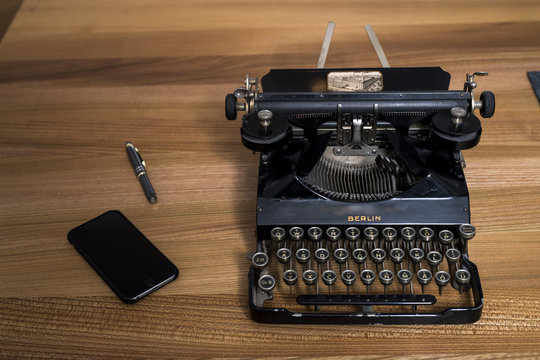 Eine alte deutsche Schreibmaschine  neben einem Kugelschreiber und einem Modernen Smartphone auf einem Holztisch