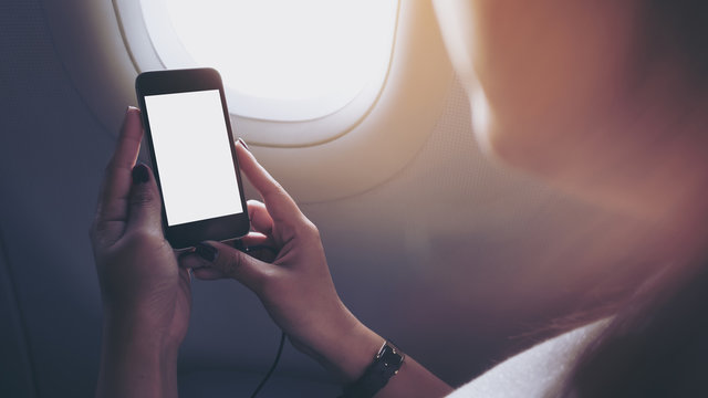 Mockup image of a woman holding and looking at black smart phone with black white screen next to an airplane window with clouds and sky background