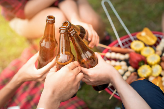 Group of friends clinking bottle of beer during camping outdoor party with barbecue in background