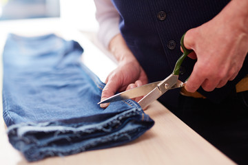 Closeup of tailors hands cutting jeans pants with big metal scissors on table in atelier