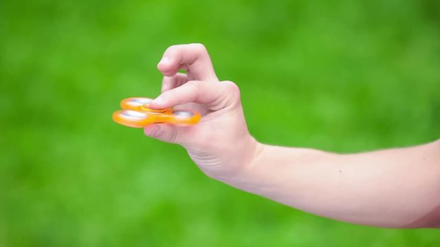 Male child hand holding popular fidget spinner toy - close up, outdoors. Boy playing with a orange Spinner in park.
