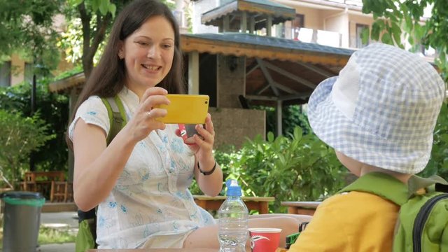 Mother takes a picture of a cute kid in the garden on the phone. Young woman makes some photo on a yellow smartphone