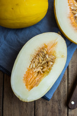 Ripe organic yellow melons, halved and whole on dark plank wood background, pulp and seeds, blue linen napkin, knife, top view, rustic
